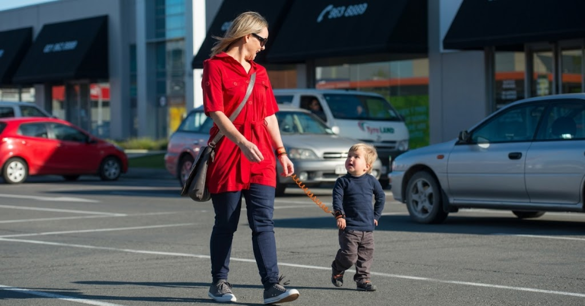 A woman walks through a busy parking lot with her young child, using a Moose Noose safety harness to prevent him from running off or eloping near parked cars.