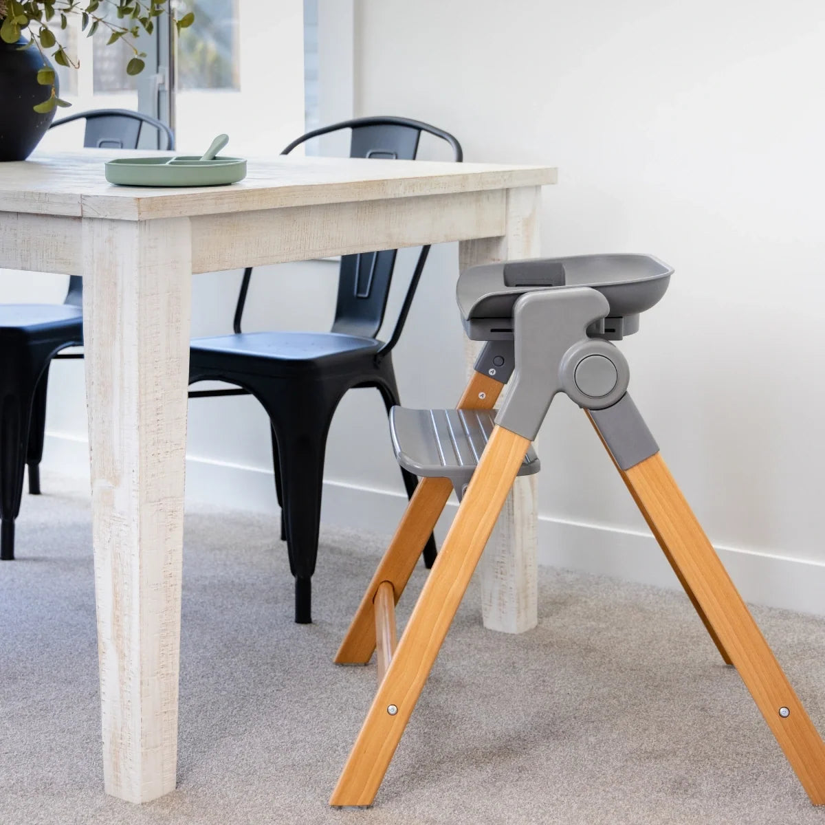 Side view of the MOOSE Frankie Foldable High Chair beside a dining table, showing wooden legs and footrest.