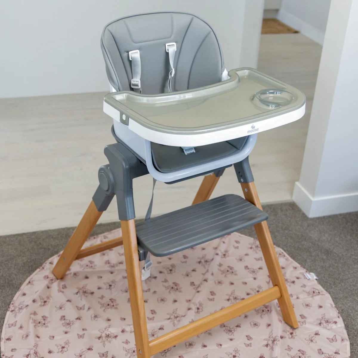 Grey MOOSE Frankie Foldable High Chair on a round rug, showing the feeding tray, wooden legs and footrest.