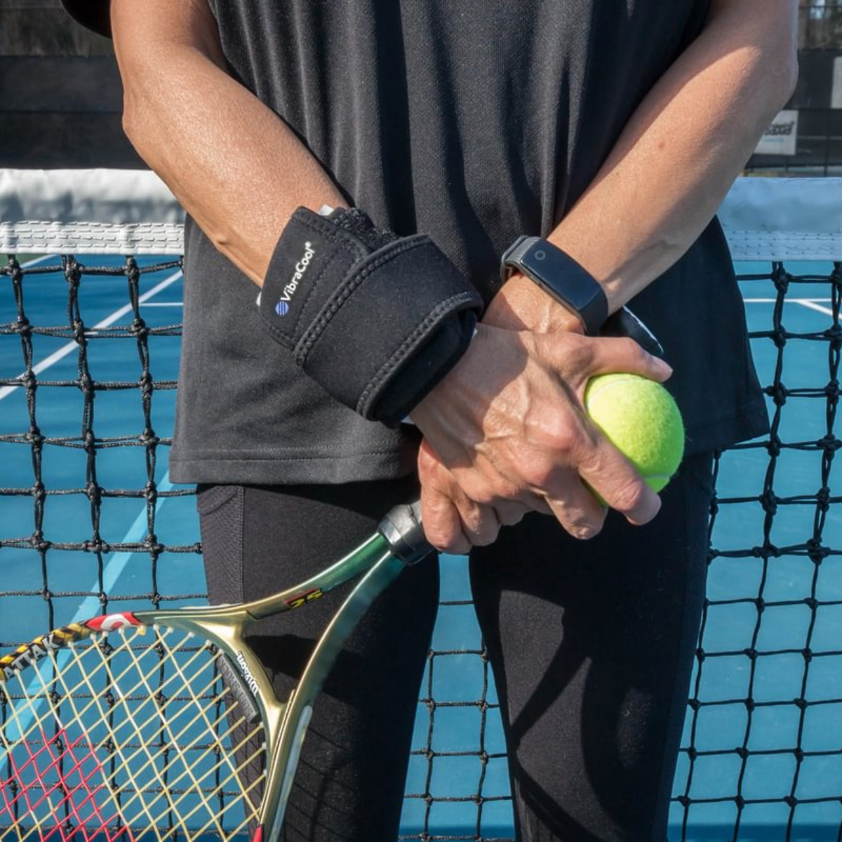 A close-up of a person on a tennis court wearing a VibraCool Eays Fit on one wrist and holding a tennis racket and tennis ball. The brace has a white logo that reads 'TENSER'.