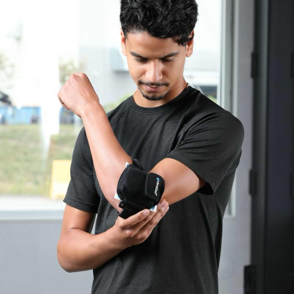 A man wearing a black athletic t-shirt demonstrates the VibraCool Easy Fit elbow support brace on his forearm in an indoor setting.