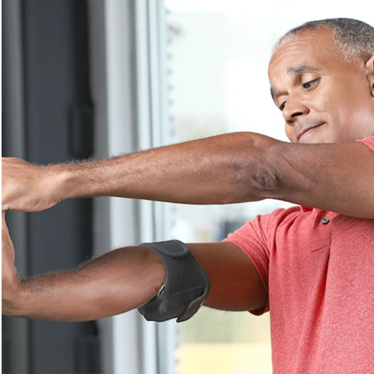 A man wearing a red polo shirt demonstrates the use of a VibraCool easy Fit for elbow support brace on his forearm, with a window in the background.