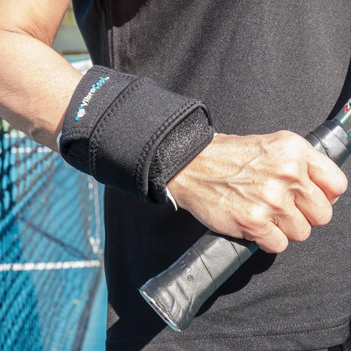 A close-up of a person's hand gripping a tennis racket handle while wearing a VibraCool East Fit ,wrist support sleeve, set against a tennis court background.